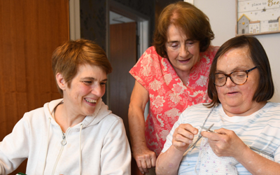 Two people sitting and one person standing around a table. One person is knitting and the other two are watching and smiling.