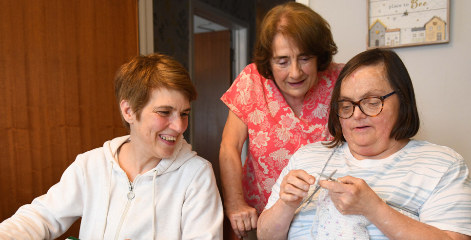 Two people sitting and one person standing around a table. One person is knitting and the other two are watching and smiling.