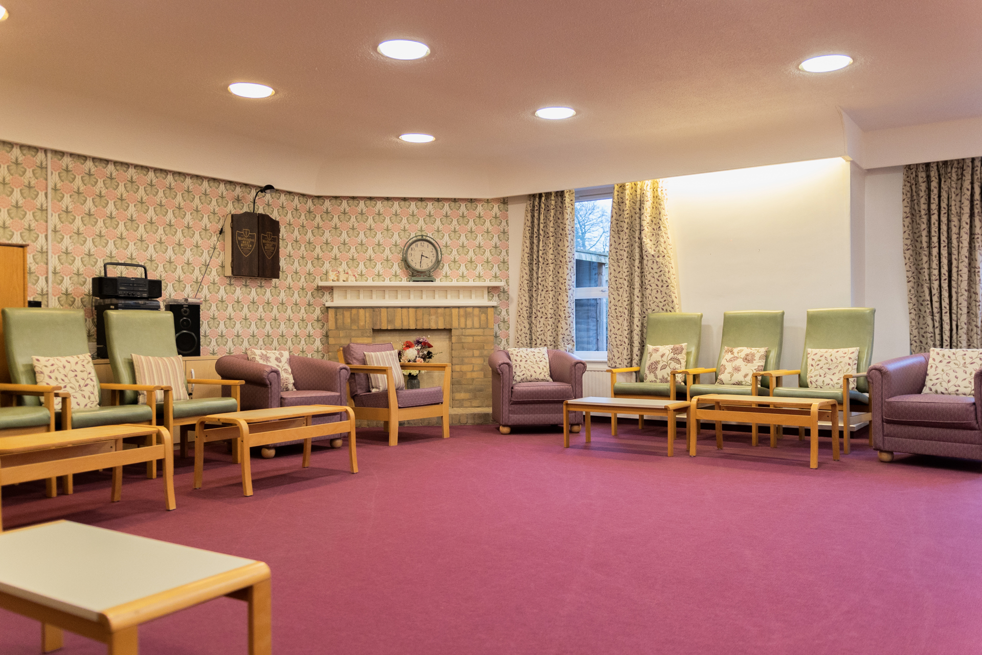 A brightly lit room with a dark pink carpet, patterned wallpaper and curtains, and armchairs set around the edge of the room.