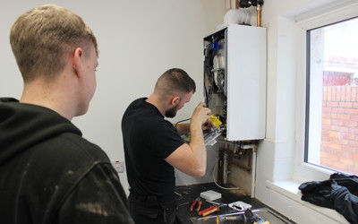 Two men in workwear are servicing a wall-mounted boiler. One man is actively working on the open boiler using tools, while the other observes closely. Various tools are scattered on the countertop below, and a window to the right lets in natural light, showing a red brick wall outside