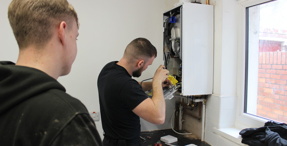 Two men in workwear are servicing a wall-mounted boiler. One man is actively working on the open boiler using tools, while the other observes closely. Various tools are scattered on the countertop below, and a window to the right lets in natural light, showing a red brick wall outside