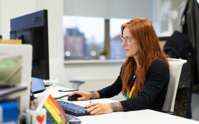 A woman with long red hair and glasses working at a desk in an office, focused on her computer screen. She is wearing a black top, a rainbow lanyard, and has tattoos on her arms. On the desk are a keyboard, mouse, and a mug with a rainbow design that says 'I ❤️'. A rainbow pride flag is also visible, along with other office supplies. A window in the background shows an urban cityscape
