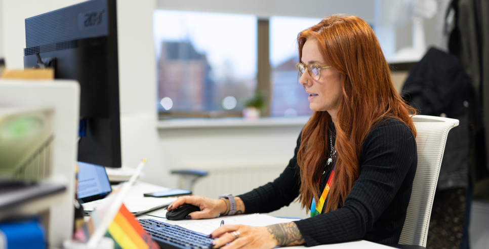 A woman with long red hair and glasses working at a desk in an office, focused on her computer screen. She is wearing a black top, a rainbow lanyard, and has tattoos on her arms. On the desk are a keyboard, mouse, and a mug with a rainbow design that says 'I ❤️'. A rainbow pride flag is also visible, along with other office supplies. A window in the background shows an urban cityscape
