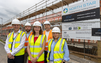 A group of people wearing hard hats and high vis vests stand outside an active building site smiling