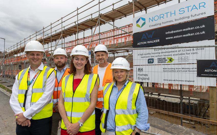 A group of people wearing hard hats and high vis vests stand outside an active building site smiling