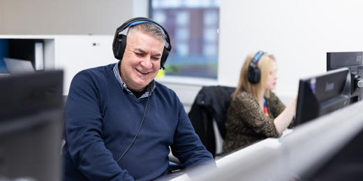 A man sat at a desk in front of a computer smiling with a headset on. 
