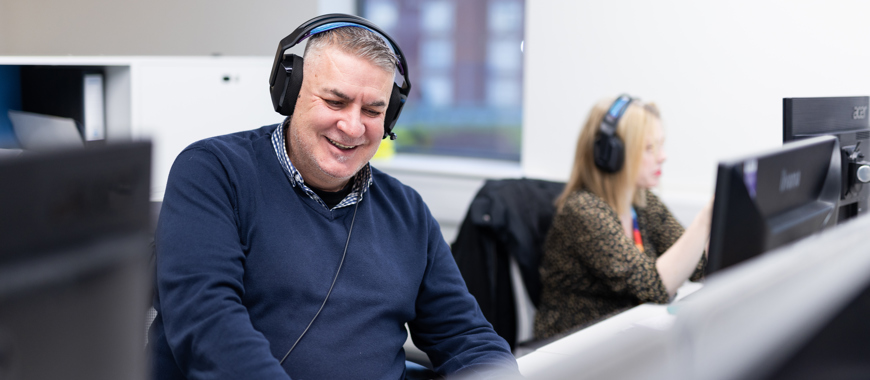 A man sat at a desk in front of a computer smiling with a headset on. 