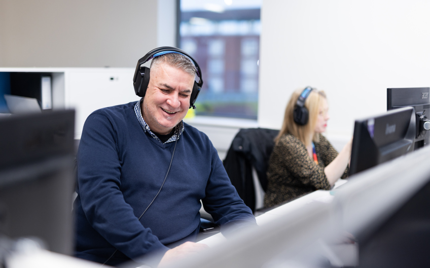 A man sat at a desk in front of a computer smiling with a headset on. 