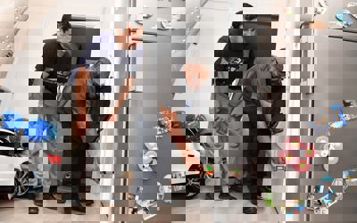 A housing officer uses a handheld device to inspect a wall inside a home, while the resident watches closely. They are in a kitchen area with a fridge covered in magnets and children’s toys visible in the background.  Let me know if you want a version tailored for accessibility compliance or a shorter version for web use.