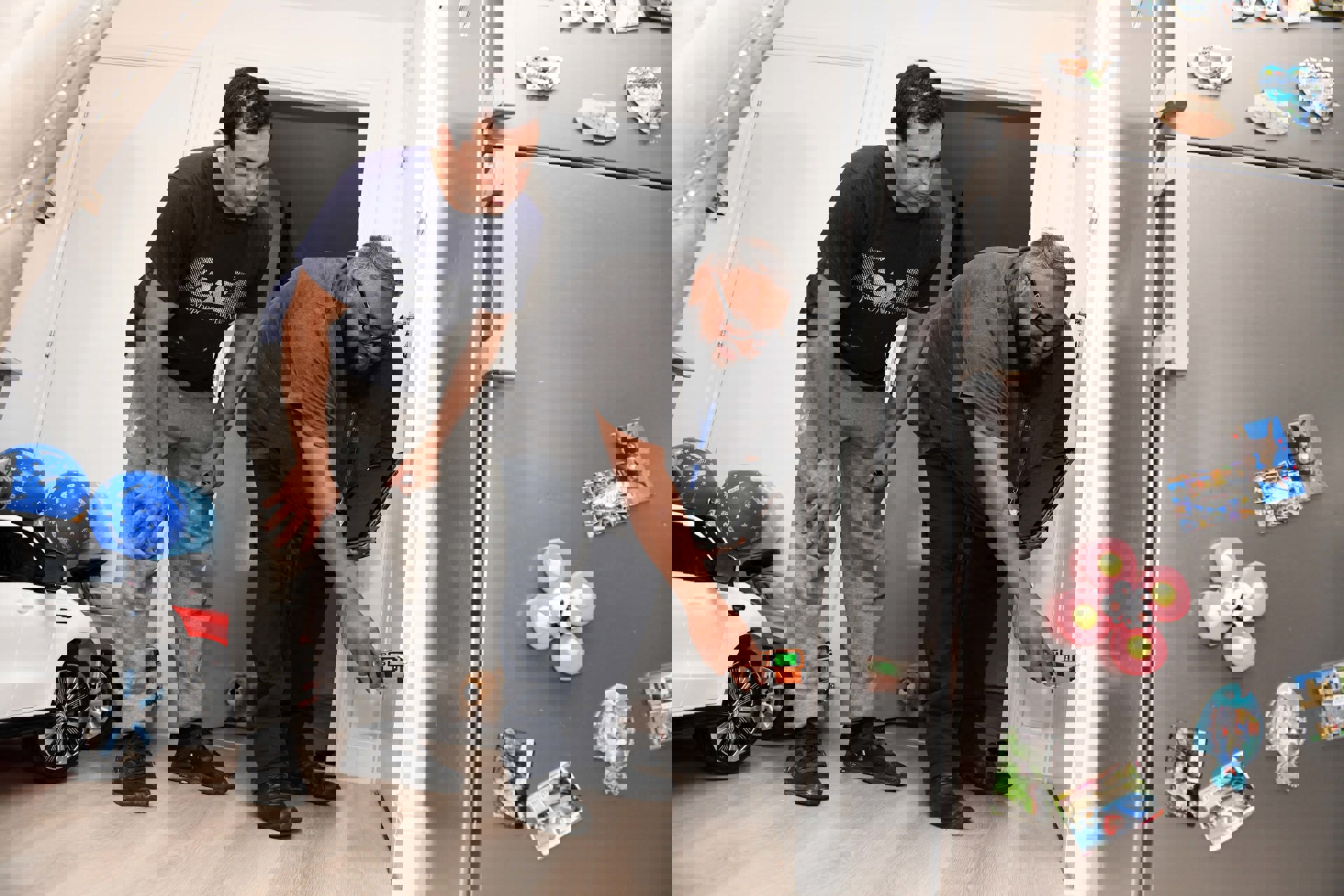 A housing officer uses a handheld device to inspect a wall inside a home, while the resident watches closely. They are in a kitchen area with a fridge covered in magnets and children’s toys visible in the background.  Let me know if you want a version tailored for accessibility compliance or a shorter version for web use.