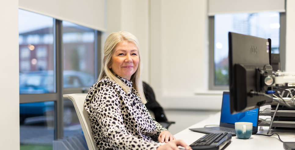 A woman with long blonde hair sits at a desk in an office, smiling at the camera. She is wearing a patterned black and white blouse and using a computer mouse. A keyboard, monitor, and a blue and white mug are on the desk in front of her. Large windows in the background show a car park and buildings outside