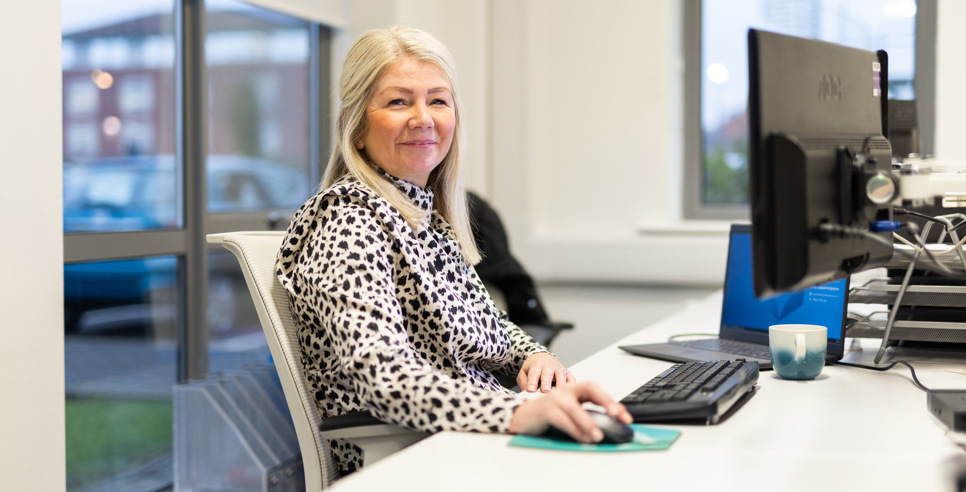 A woman with long blonde hair sits at a desk in an office, smiling at the camera. She is wearing a patterned black and white blouse and using a computer mouse. A keyboard, monitor, and a blue and white mug are on the desk in front of her. Large windows in the background show a car park and buildings outside