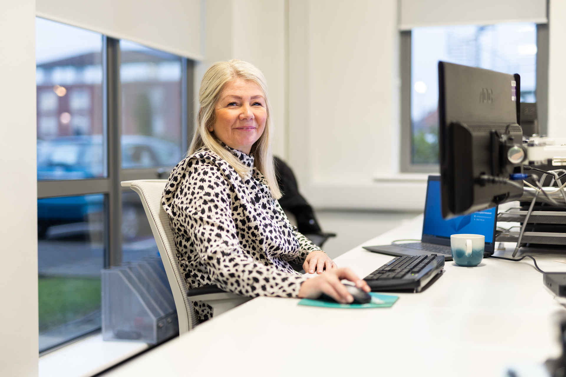 A woman with long blonde hair sits at a desk in an office, smiling at the camera. She is wearing a patterned black and white blouse and using a computer mouse. A keyboard, monitor, and a blue and white mug are on the desk in front of her. Large windows in the background show a car park and buildings outside