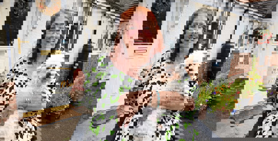 A smiling woman sitting in a wheelchair outside her home, holding a fluffy white dog on her lap and a large mug that reads 'Magnificent Mum.' She is wearing a white dress with green and black spots. Behind her is a white front door with a floral wreath and a garden with colorful potted flowers.